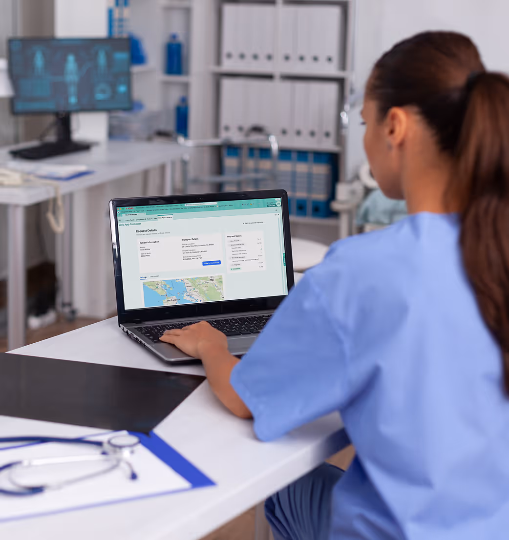 Healthcare worker in blue scrubs using a laptop showing patient transport details in a medical office.