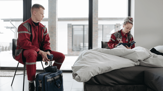 Two community paramedics in red uniforms check on a patient at home, one taking notes beside the bed.