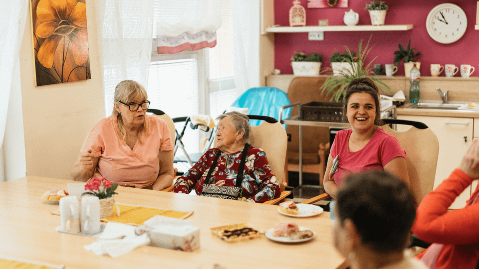 Older adults and caregivers chat around a table in a bright day center, the kind of setting PACE programs serve.